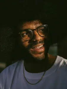 A person with a large afro, wearing round glasses, a white shirt, and a gold chain, looks slightly to the side in natural light.